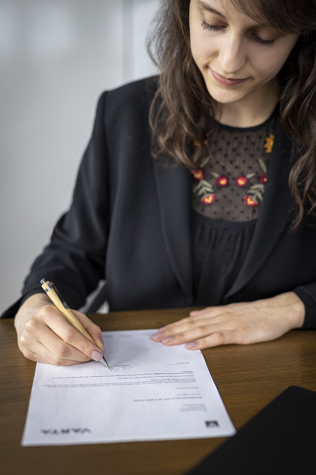 Woman with long brown hair in black blazer signing a document with a wooden pen at a desk