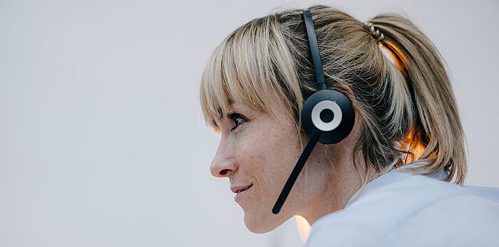 Side profile of a smiling blonde woman with ponytail and bangs wearing a black headset with microphone