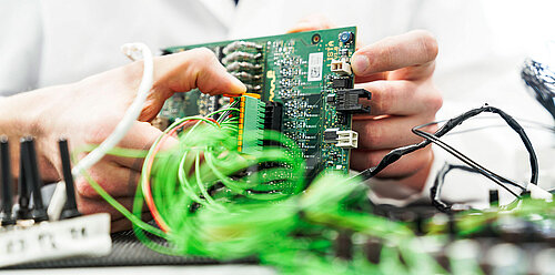 Close-up of hands holding a green circuit board connected to bundles of green wires in a lab setting