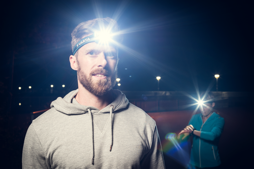 Bearded man in grey hoodie wearing bright Varta headlamp at night, woman in teal jacket checks watch in background