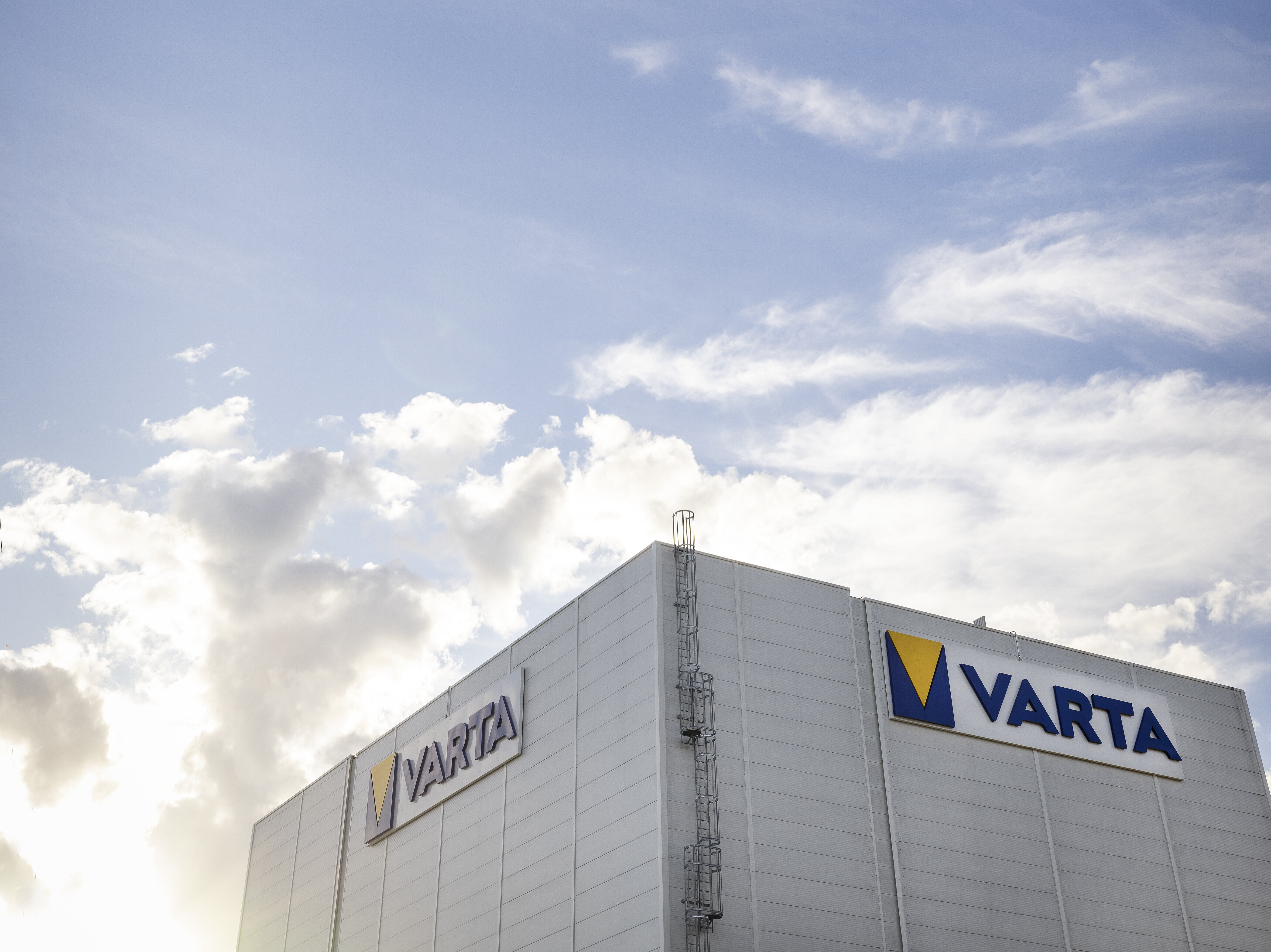 Modernes Industriegebäude mit zwei VARTA-Logos an der Fassade und Außenleiter vor blauem Himmel mit Wolken