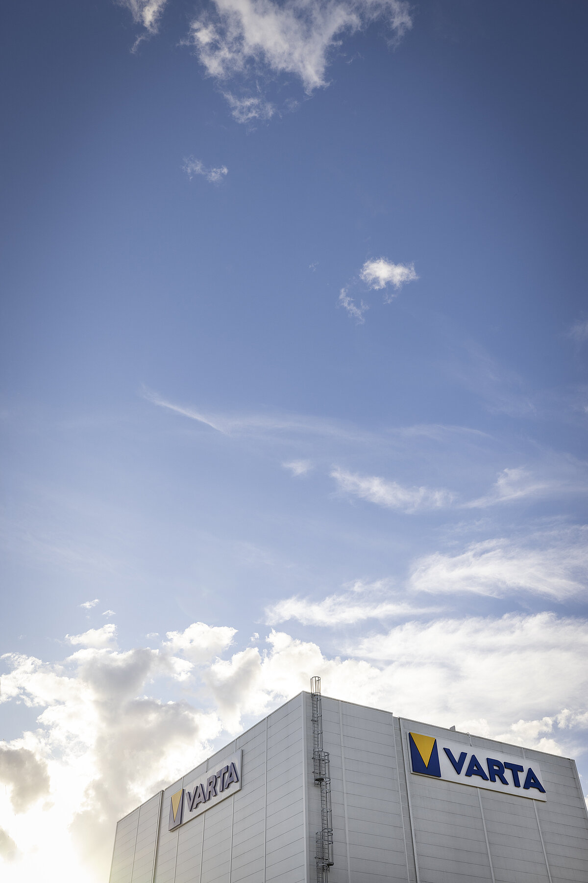 Modern industrial building with two VARTA logos on its facade, set against a bright sky with scattered clouds