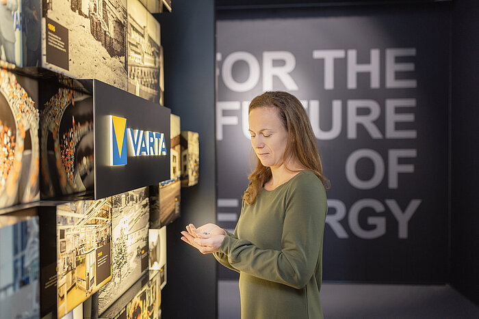 Woman in green top examines small object beside VARTA logo display and "For the Future of Energy" wall in exhibition space