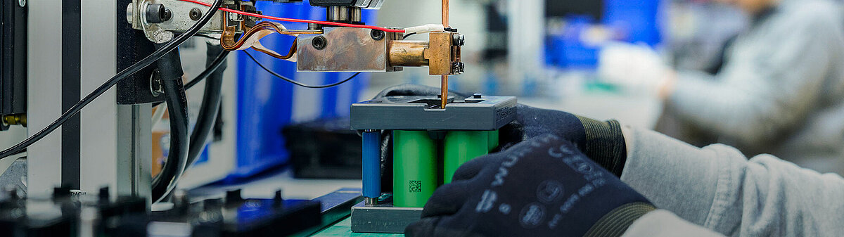 Gloved hand positioning green lithium-ion battery cells under a spot welding machine in an industrial setting