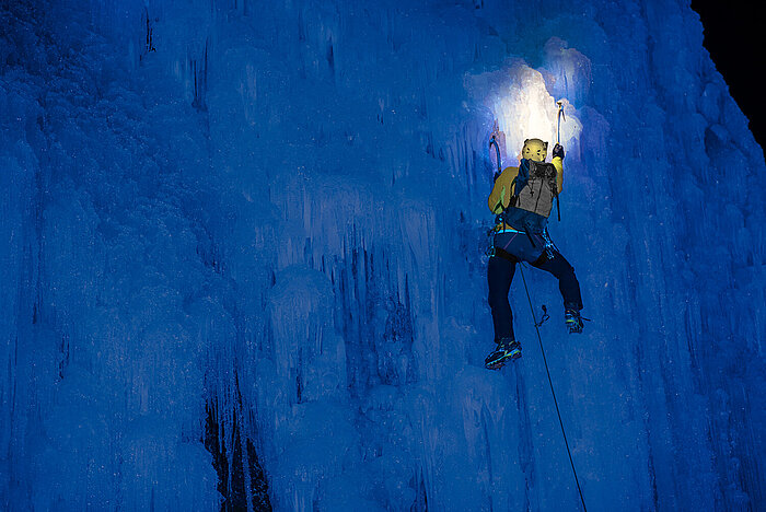 Ice climber in yellow jacket with helmet lamp ascending a massive blue frozen waterfall at night, ropes and ice axes visible
