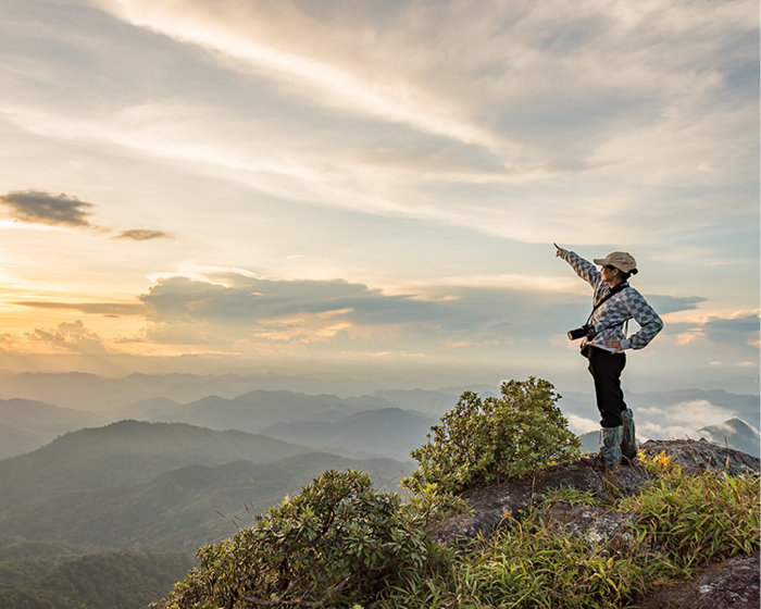 Hiker with camera stands on mountain peak, pointing toward distant misty hills under cloudy sky at sunset