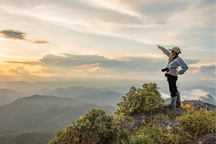 Hiker with camera stands on mountain peak, pointing toward distant misty hills under cloudy sky at sunset