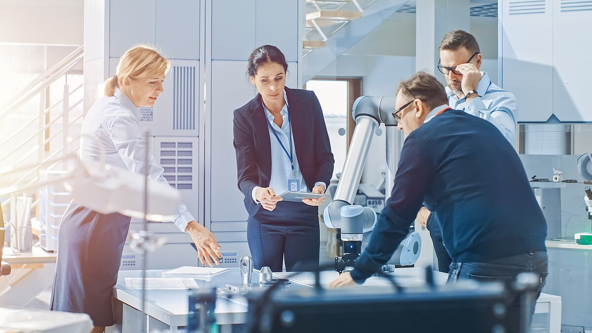 Four professionals gathered around a robotic arm in a bright industrial lab, one holding a tablet during discussion