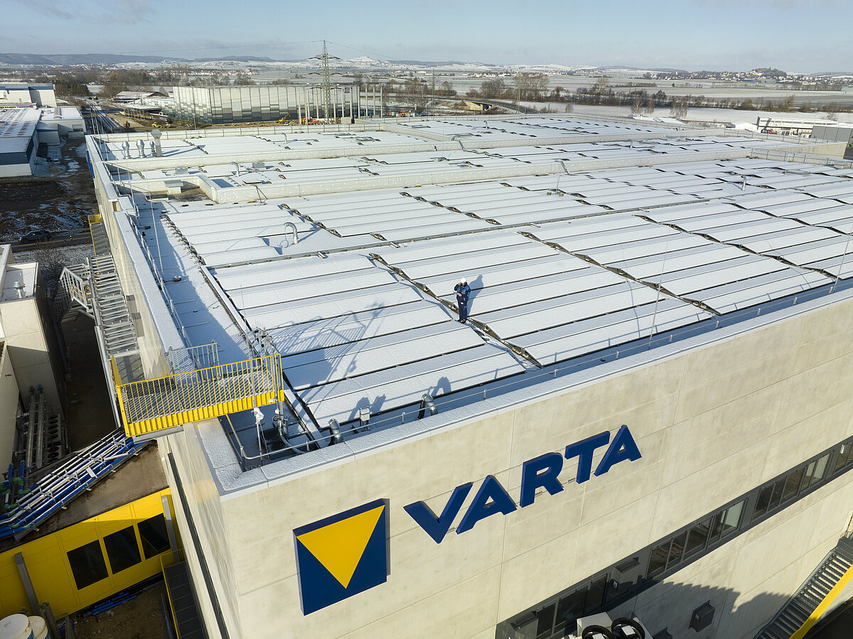 Aerial view of a VARTA factory building with snow-covered roof panels and a worker in blue standing on top