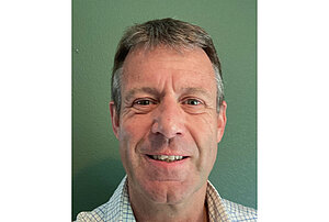 Smiling middle-aged man with short graying hair, wearing a checkered shirt, against a dark green wall