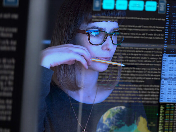 Woman with glasses and bangs holds pencil to lips, studying transparent screen with data, text and stock market figures