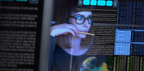 Woman with glasses and bangs holds pencil to lips, studying transparent screen with data, text and stock market figures