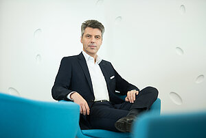 Man in dark suit and white shirt sits relaxed on a blue sofa, looking at the camera against a white wall