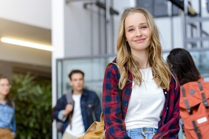 Smiling young woman with long blonde hair in plaid shirt stands in building entrance, people blurred in background