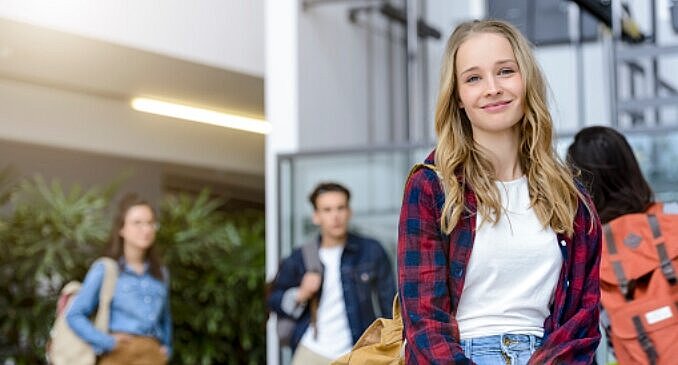 Smiling young woman with long blonde hair in plaid shirt stands in building entrance, people blurred in background