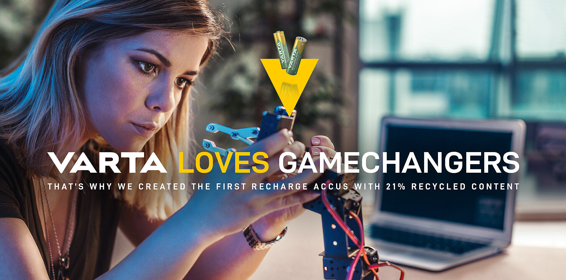 Mladá žena pracuje s robotickou rukou, reklama VARTA s nápisem „Varta loves gamechangers" a dvěma bateriemi