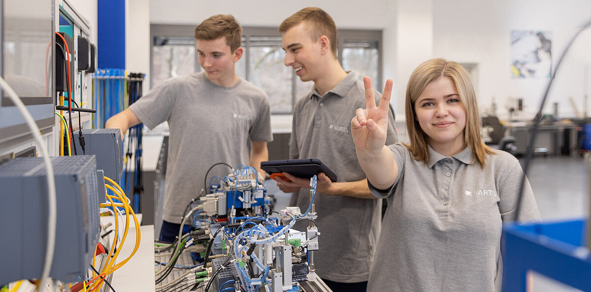 Three young trainees in grey VARTA shirts work with industrial automation equipment; woman in front makes a peace sign