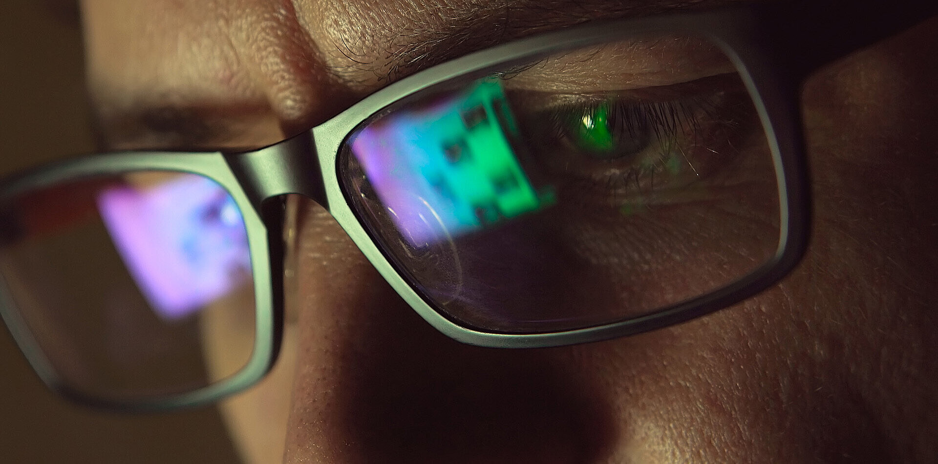 Close-up of a person's eye behind glasses, with colorful screen reflections in green and purple on the lenses
