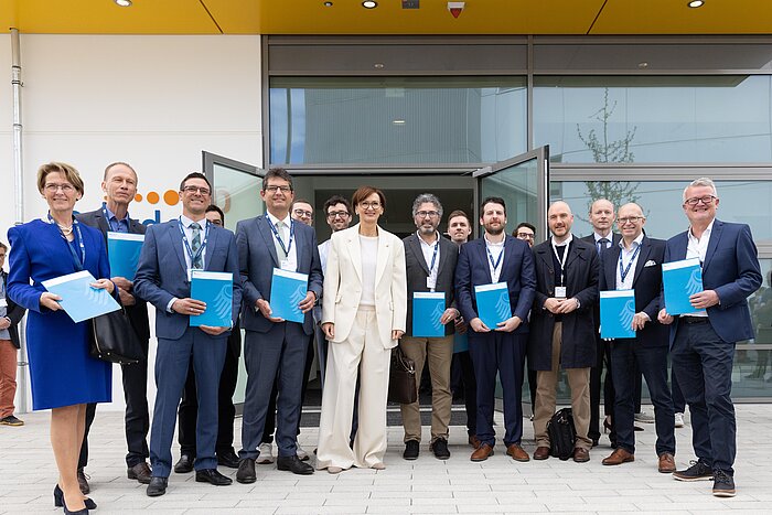 Group of formally dressed people holding blue folders posing in front of a building entrance with glass doors