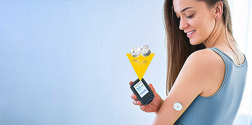 Smiling woman in blue top holds glucose meter showing 5.5 near sensor on arm, two Varta coin cell batteries above