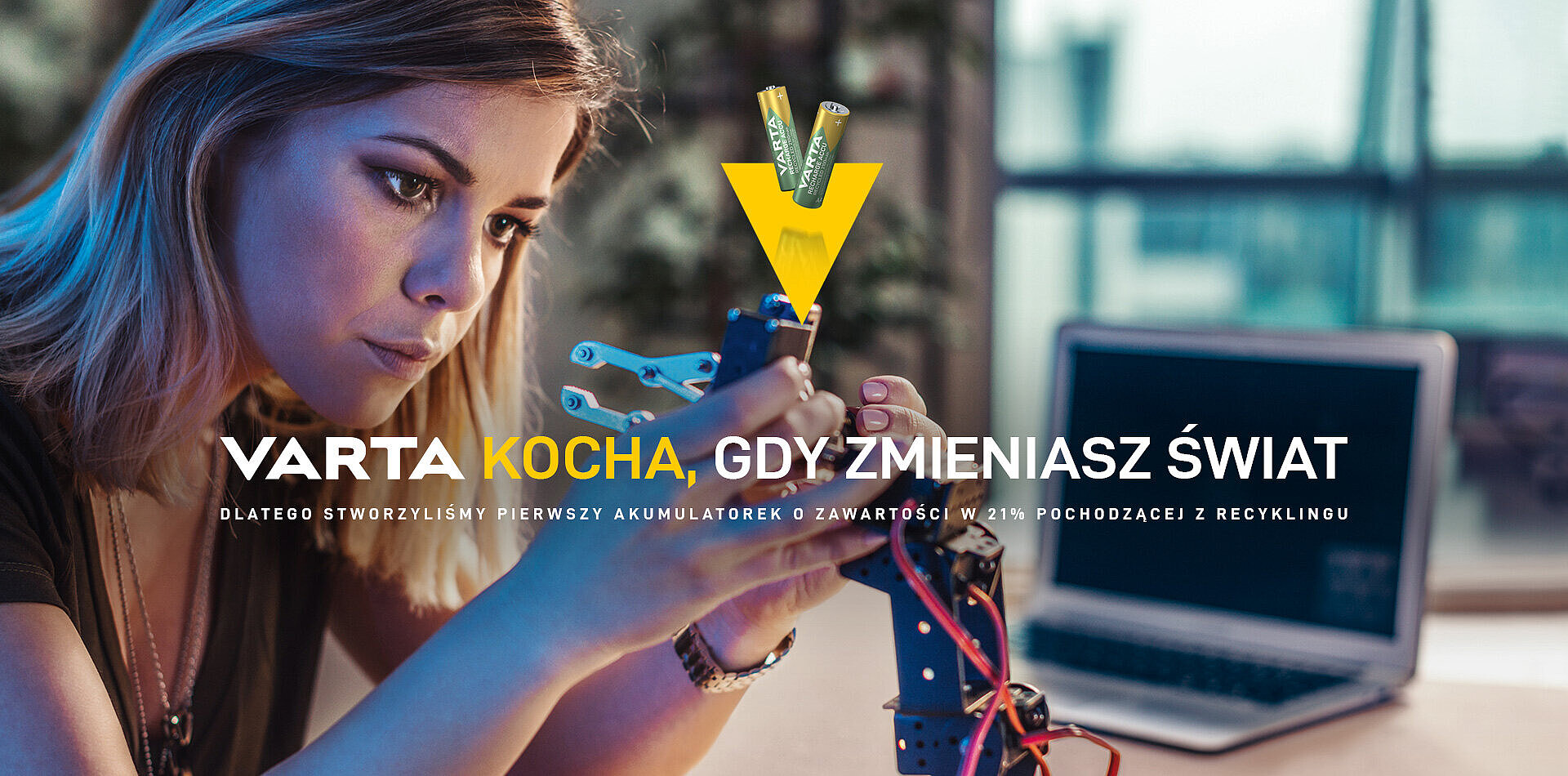 Młoda kobieta składa robota przy laptopie, obok logo VARTA z bateriami i hasłem "Varta kocha, gdy zmieniasz świat