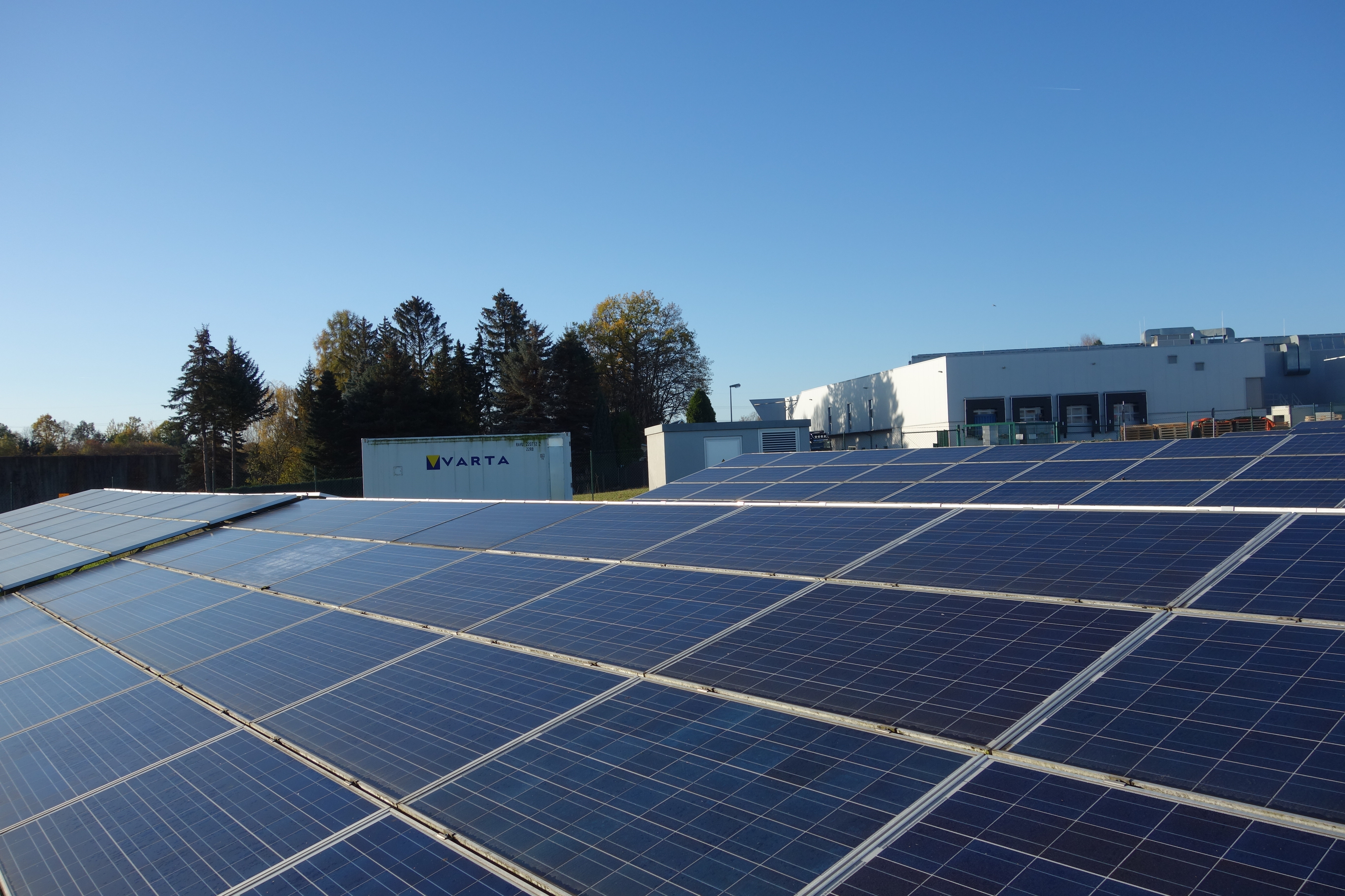 Große Photovoltaikanlage auf Flachdach, im Hintergrund Industriegebäude und VARTA-Container unter blauem Himmel