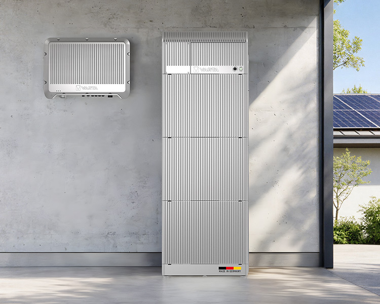 Silberner VARTA Batteriespeicher und Wechselrichter an Betonwand, rechts Hausdach mit Solarmodulen und Bäume